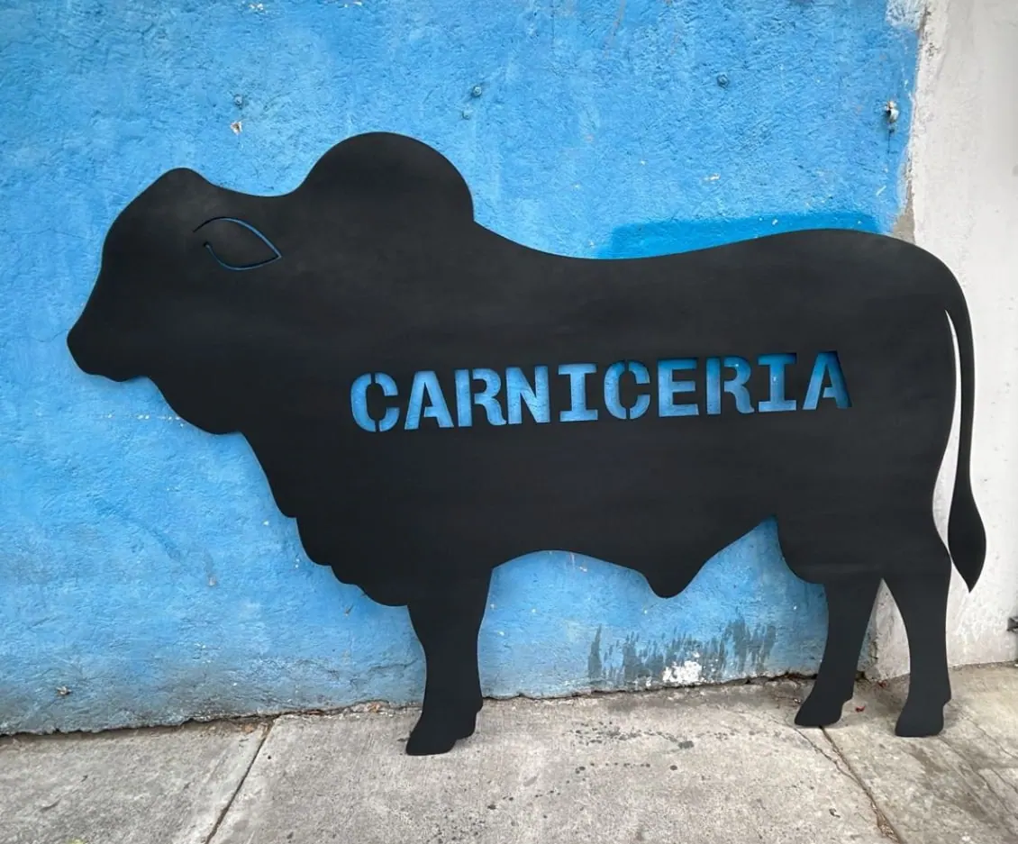Silueta de toro en lámina negra con la palabra carnicería calada, recargada en un muro azul
