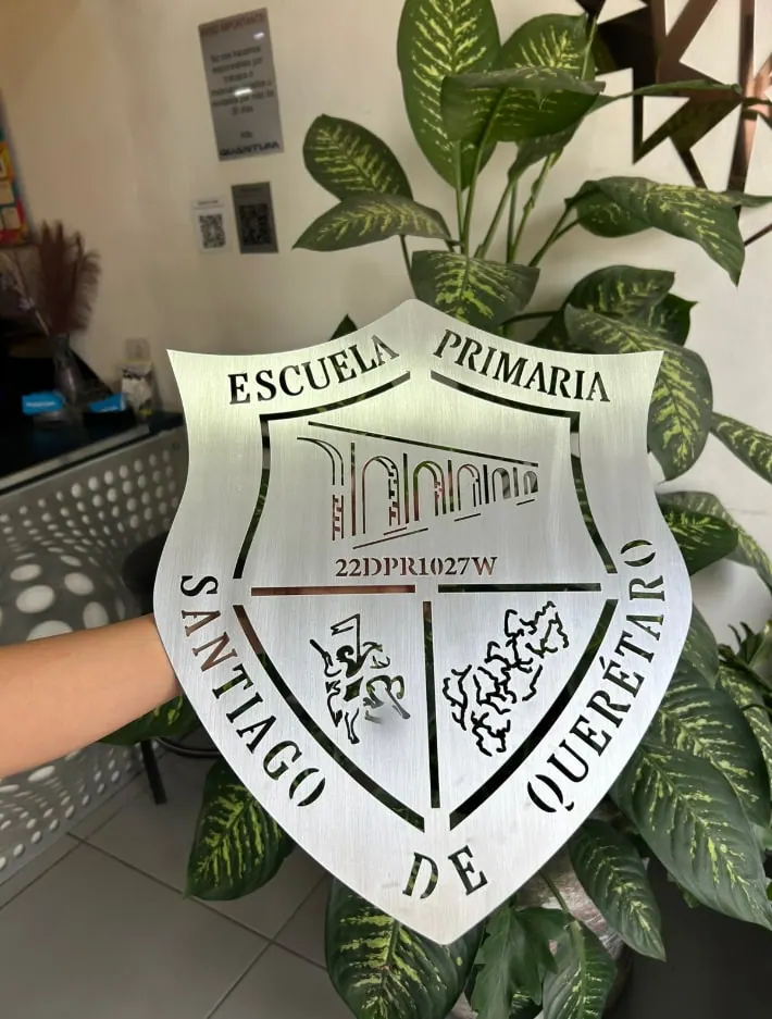 Escudo escolar calado en acero, sostenido frente a una planta